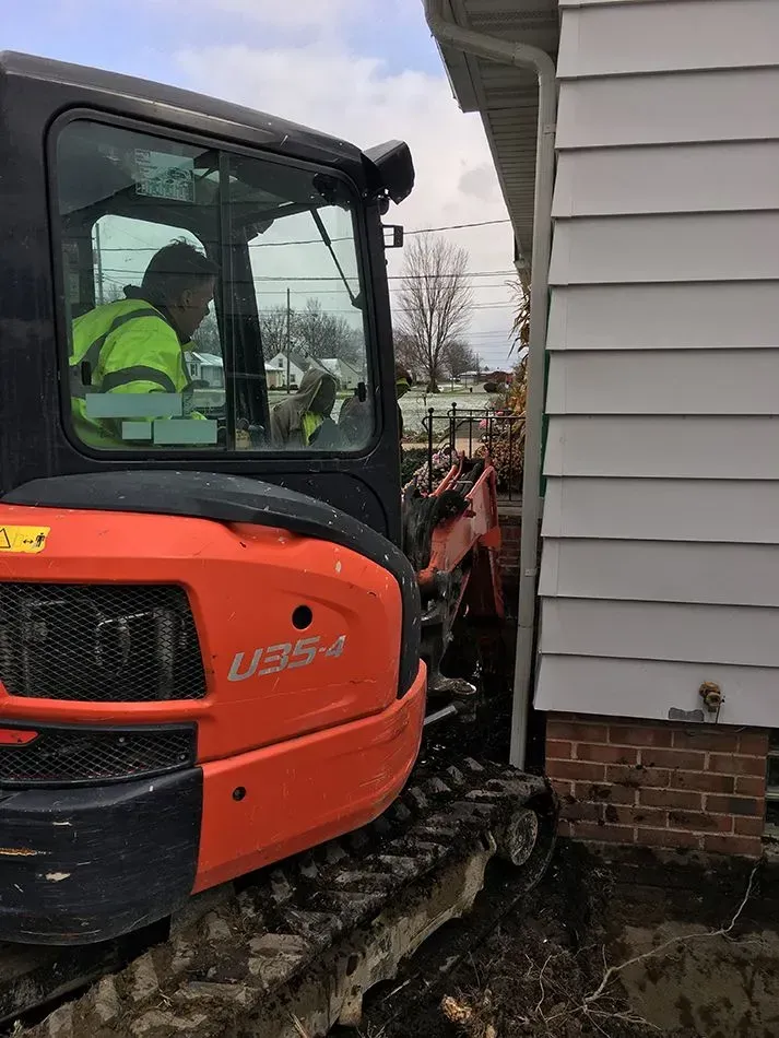 Orange excavator digging next to a white house; a person in a safety vest operates the machine.