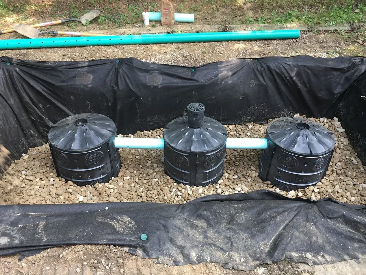 Three black septic tanks connected with pipes in a gravel-lined pit, covered with black fabric.