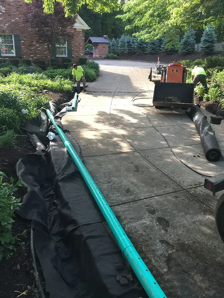Construction workers installing a drainage system along a driveway. A long blue pipe is laid out.