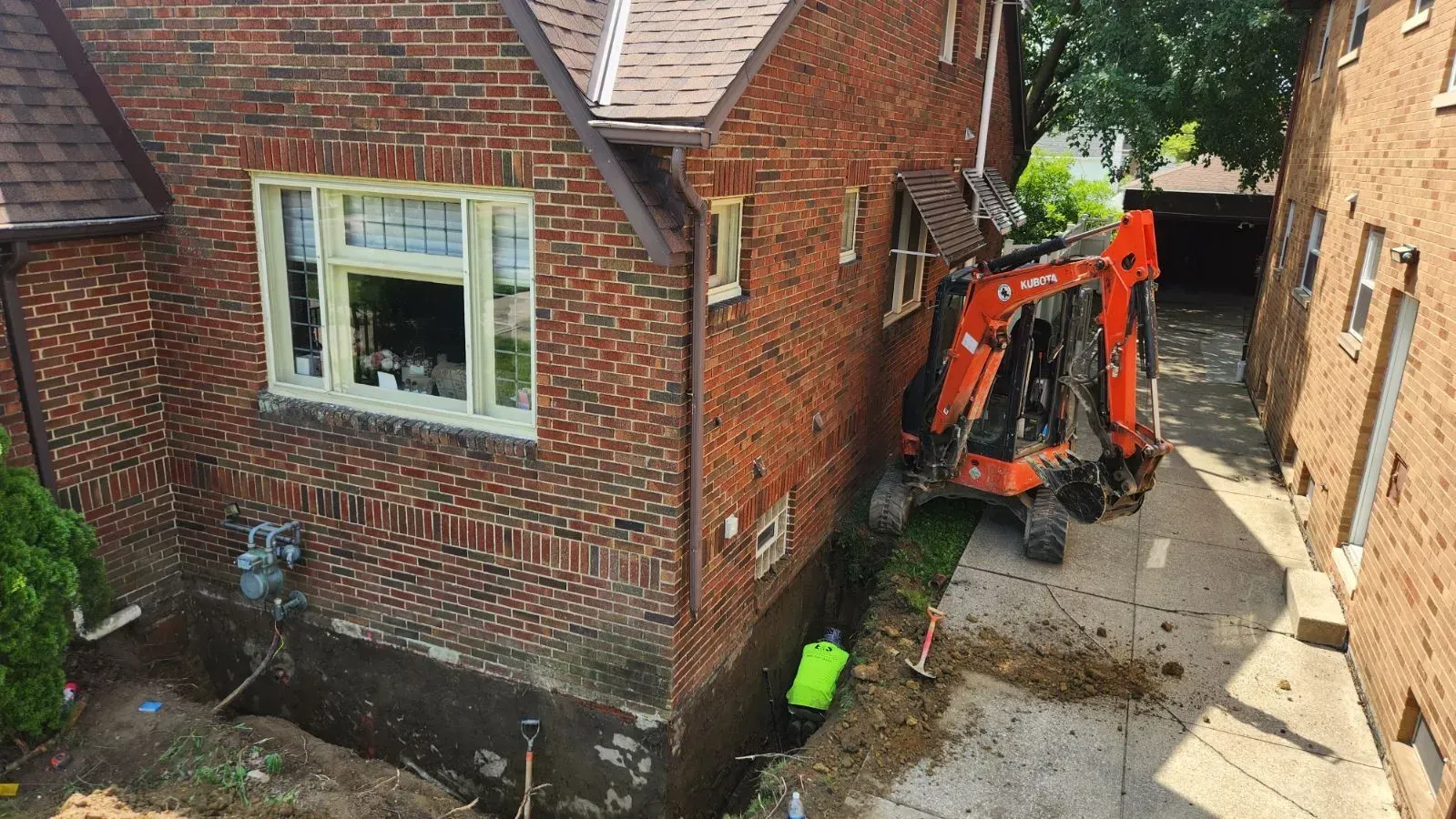 Construction: Mini-excavator digging trench beside brick house, worker in safety vest.