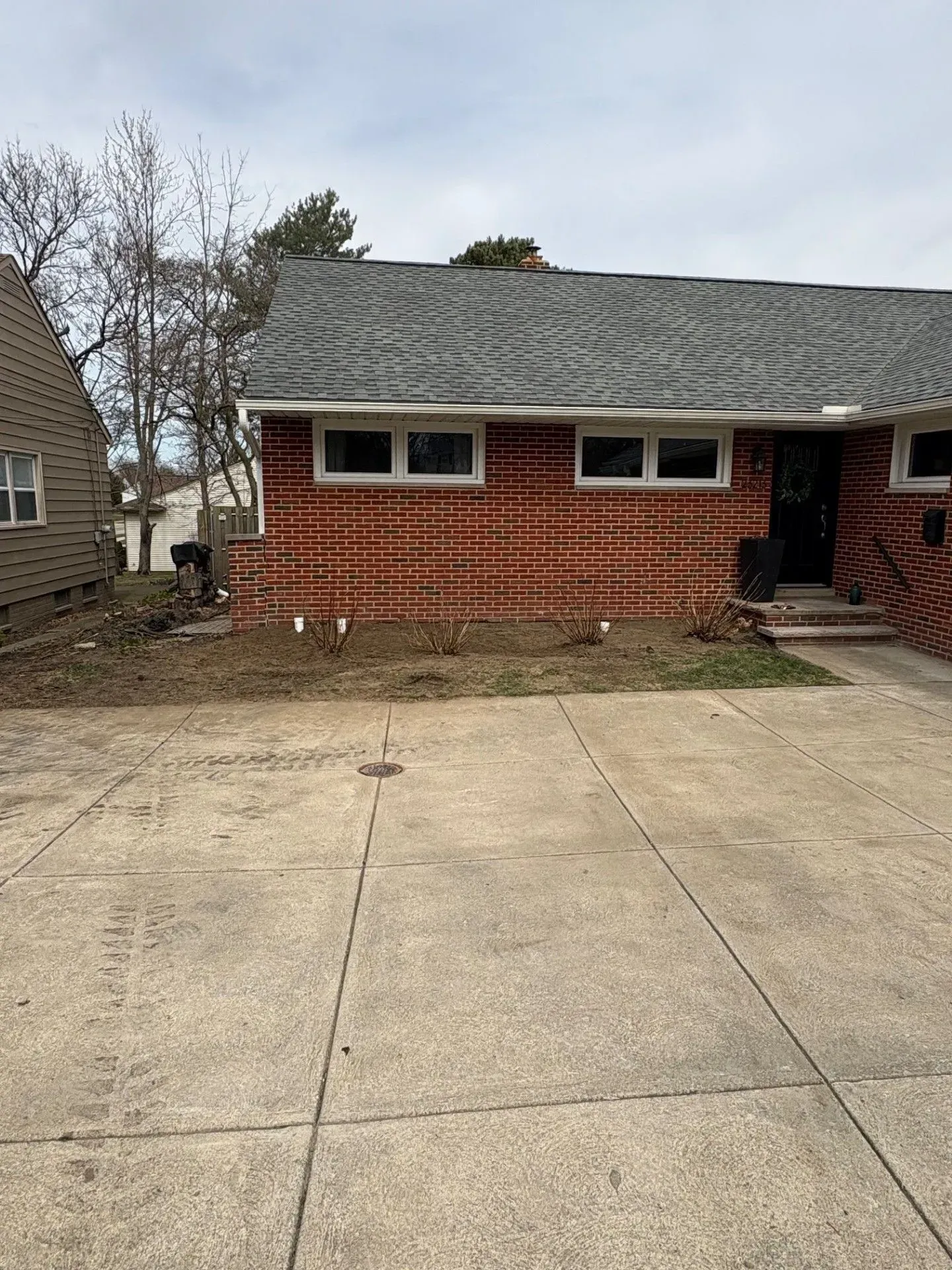 A brick house with a concrete driveway and bare patches of dirt in the front yard under a cloudy sky.