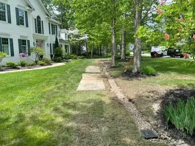 Residential front yard with lawn, stone pathway, trees, and houses.