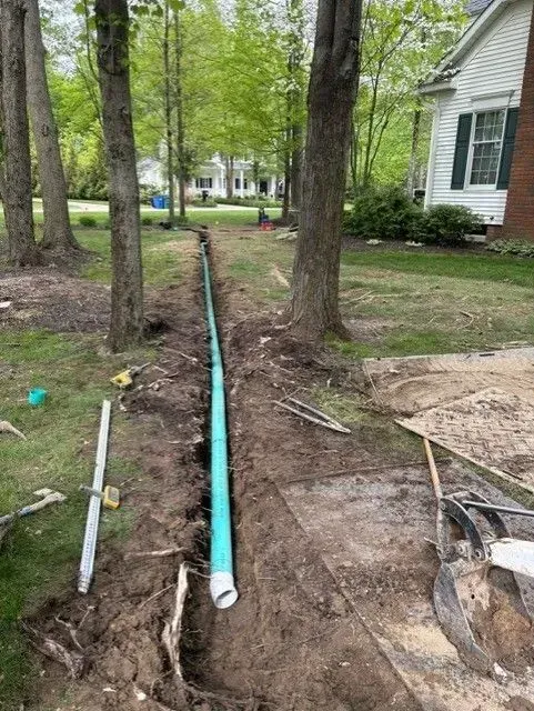 Trench dug in yard with a green pipe inside, likely for drainage, near trees and a house.