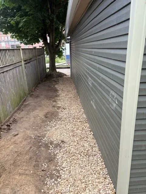 Narrow dirt path with gravel, between a gray-sided building and wooden fence, with a tree in the background.