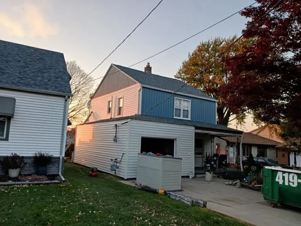 A two-story house with blue siding and a garage. A green dumpster is in the driveway, autumn trees in the background.