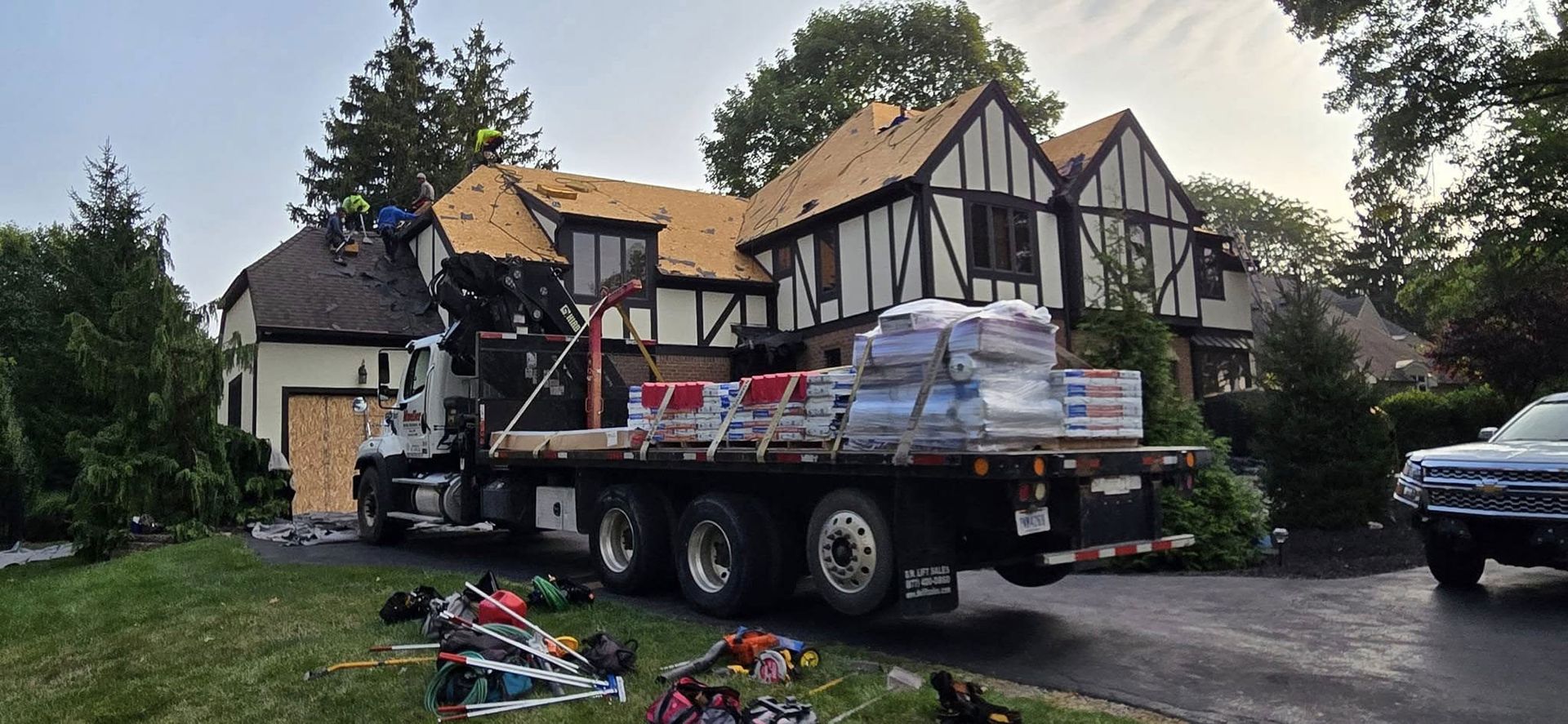 A roofing crew is working on a Tudor-style house, with a flatbed truck full of supplies parked in the driveway.