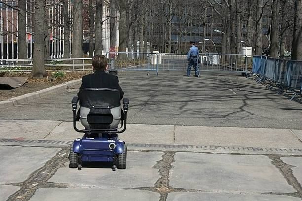 Person in blue motorized wheelchair on paved path, another person distant. Trees and barriers in background.