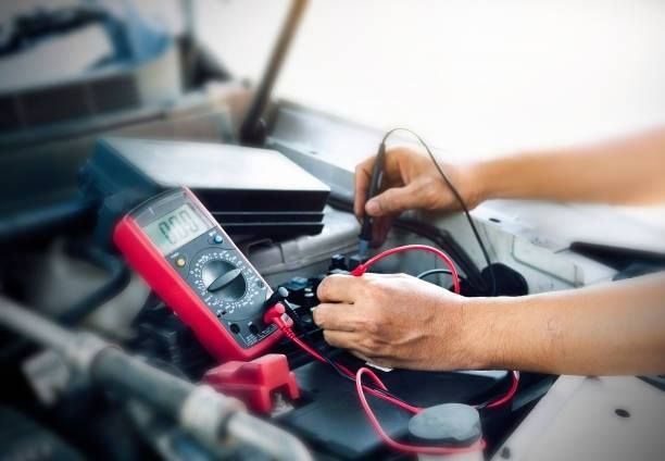 Hands using a multimeter to test a car battery under the hood. Red and black wires are connected.