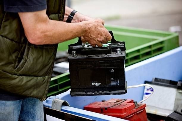 Person in vest holding a car battery, placing it in a recycling bin.
