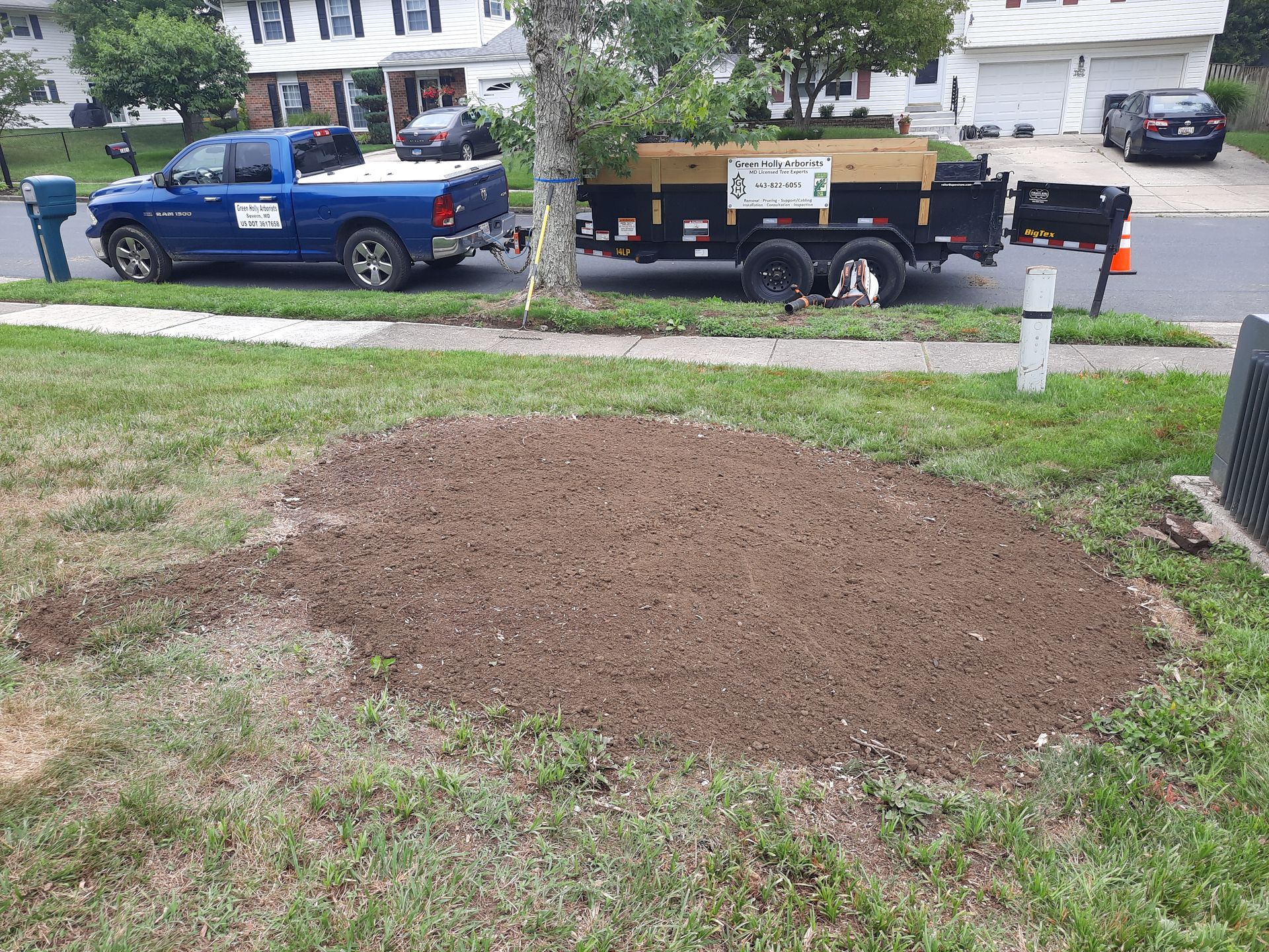Freshly turned brown soil patch in a grassy yard beside a sidewalk, with parked vehicles and houses in the background