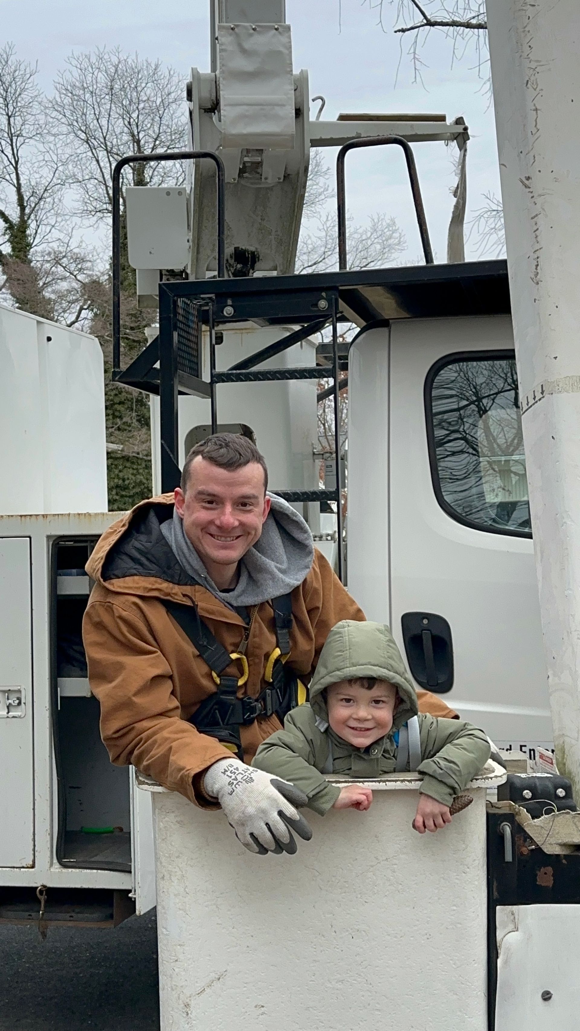 Man in brown jacket smiling beside a child in a green hat inside a white bucket lift outdoors