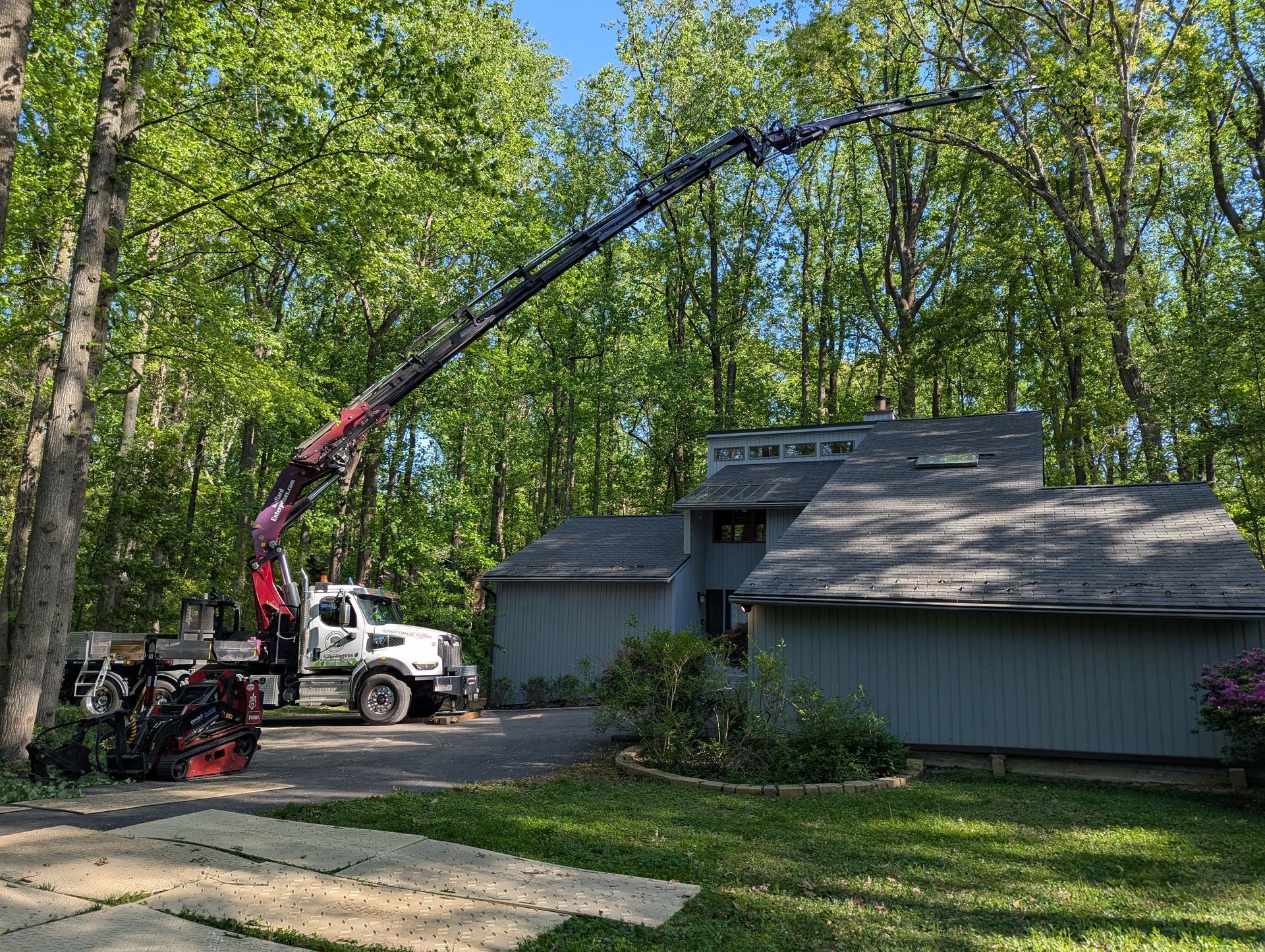 Tree-service truck with extended boom trimming branches beside a house in a wooded yard