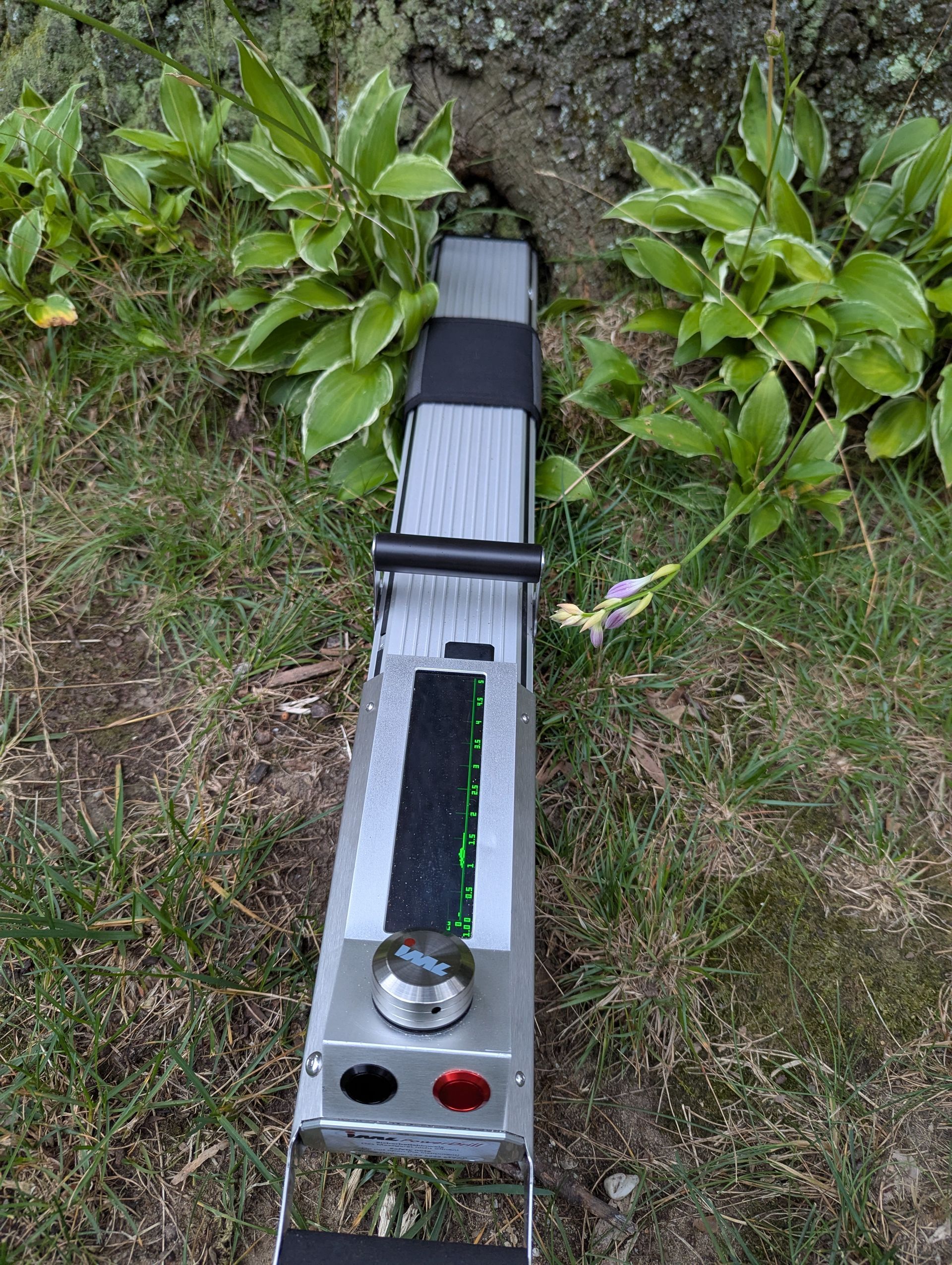 Outdoor metal device with a black handle, silver body, and red button lying on grass near plants