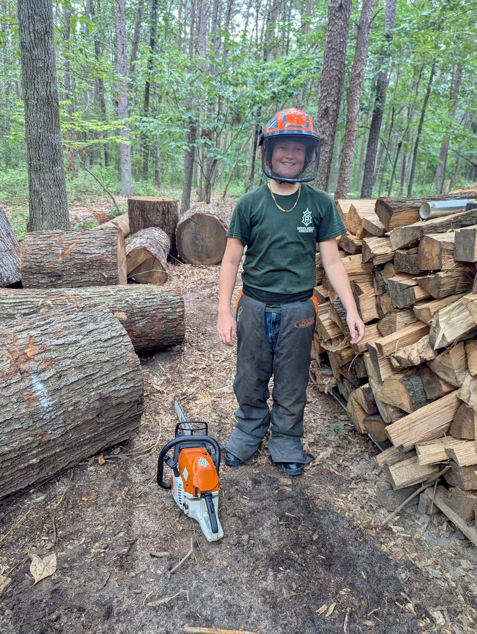 Person standing beside a chainsaw and stacked logs in a wooded area, wearing a helmet and protective gear