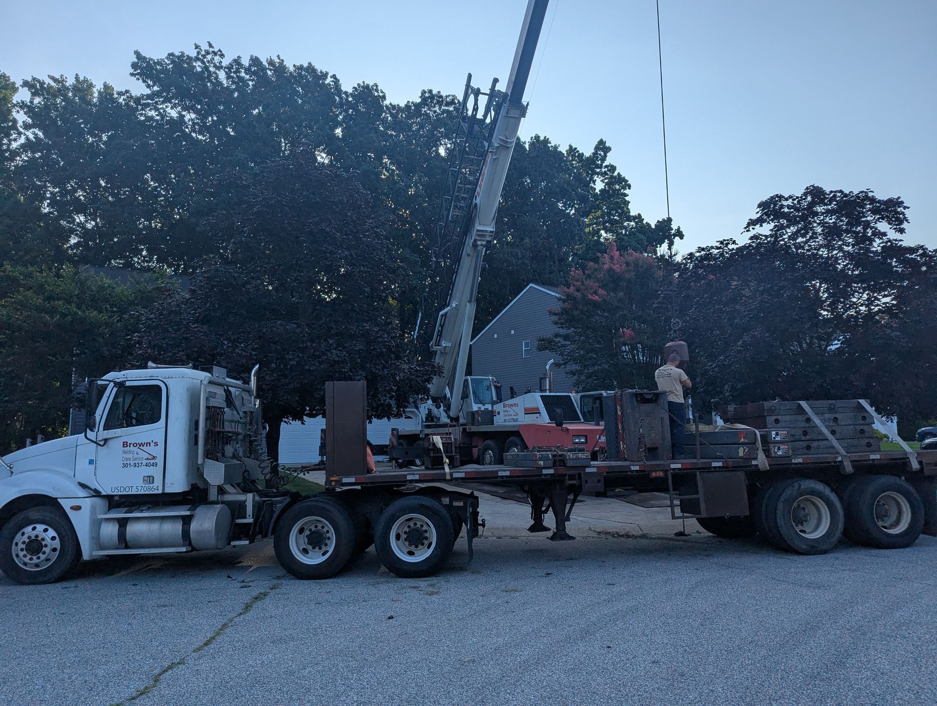 White semi truck hauling a crane on a flatbed beside trees and road work equipment