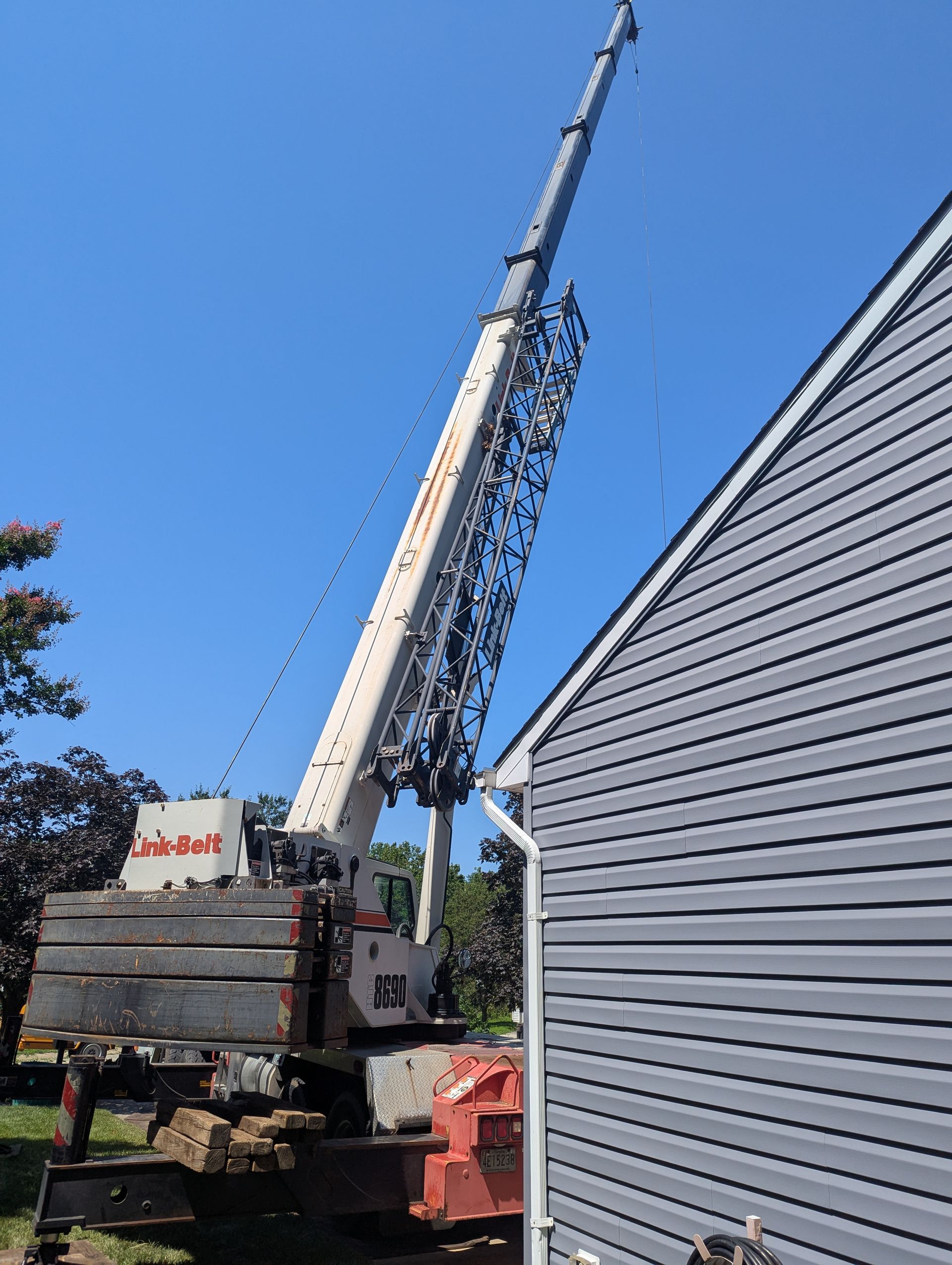 Crane lifting a tall mast beside a gray building under a clear blue sky