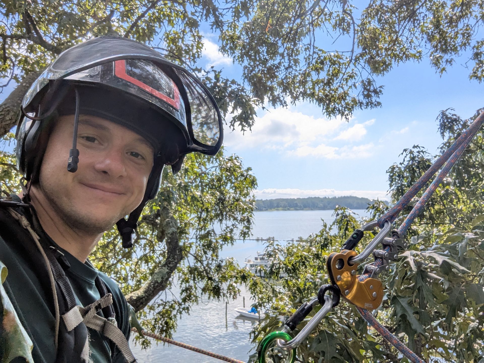 Smiling person in a helmet beside a tree, with water and a blue sky in the background.