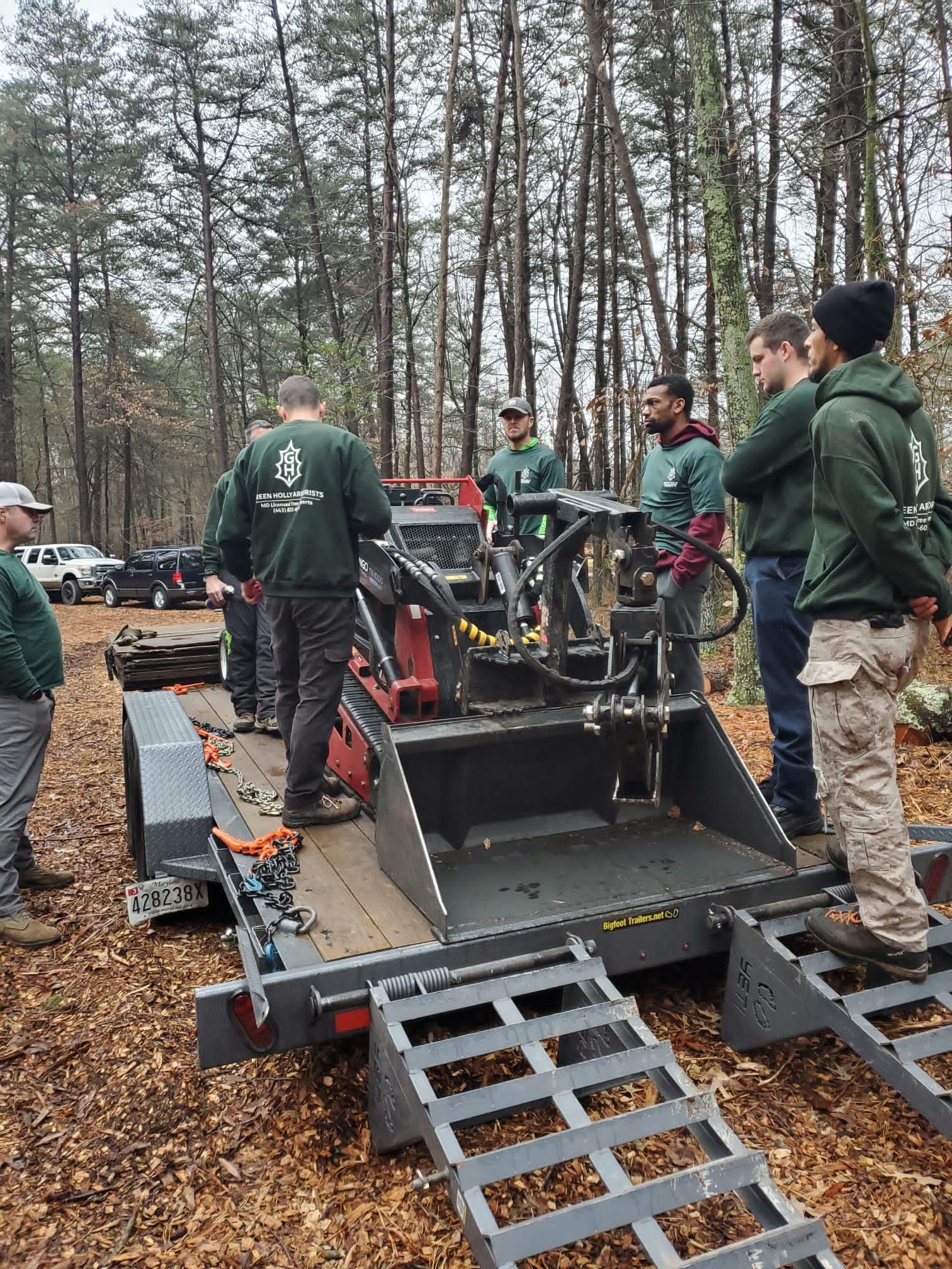 Workers in green jackets inspect a forestry machine in a wooded area with muddy ground and tall trees.