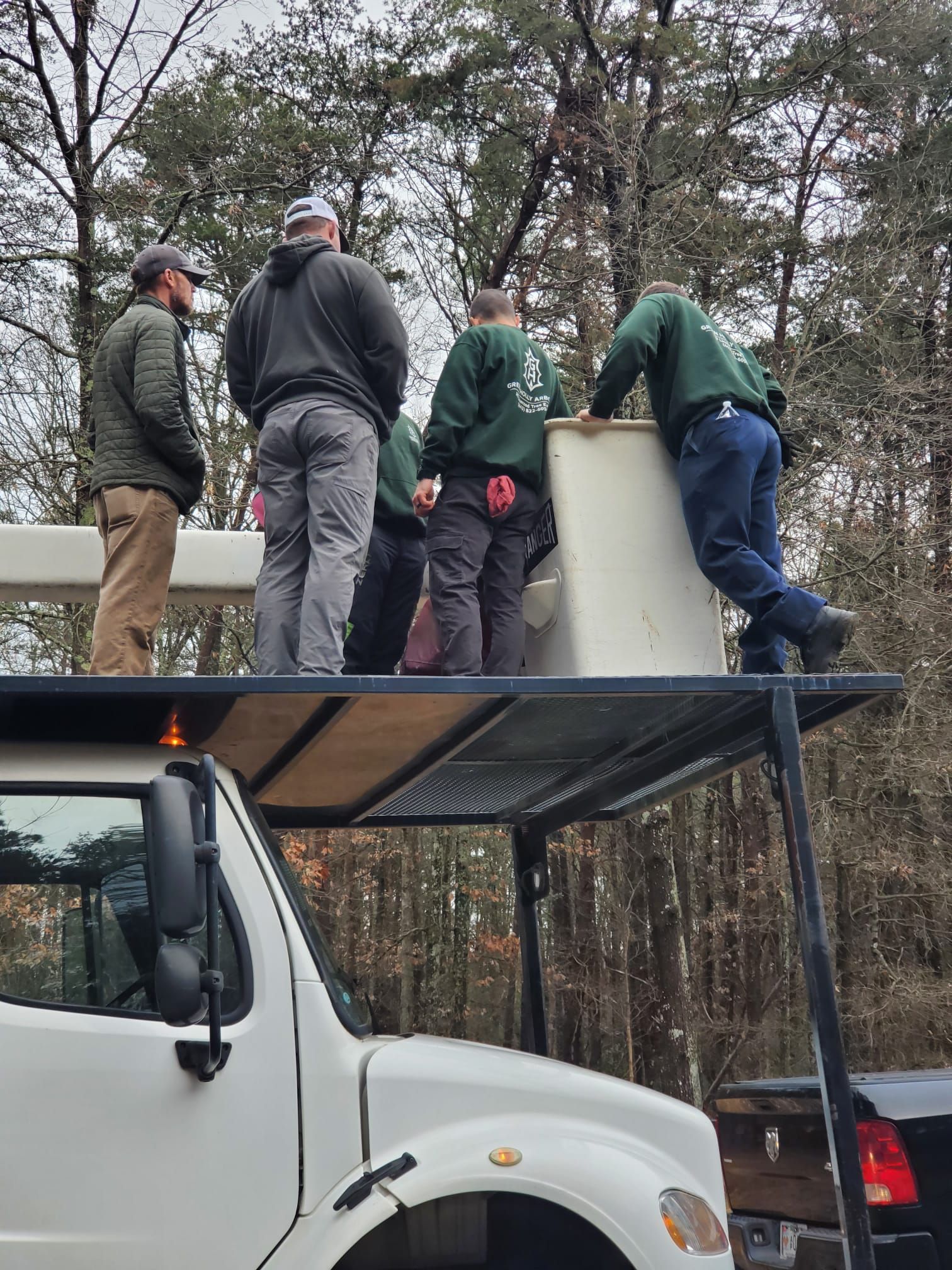 Workers on a truck platform inspecting a large white tank in a wooded outdoor area.