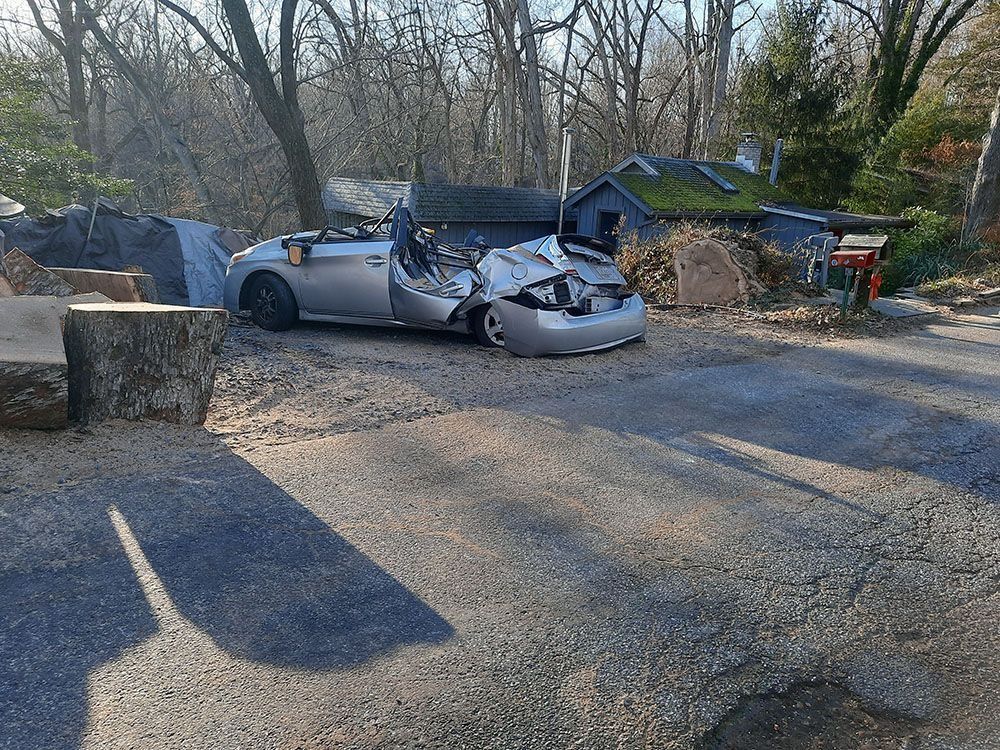 Severely damaged silver car crashed on a gravel driveway near trees and a house