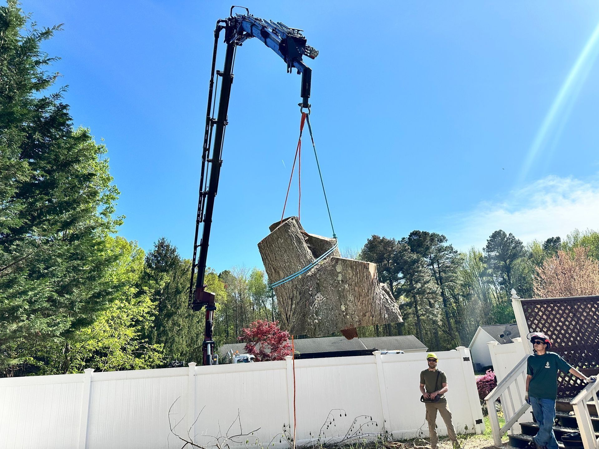 Crane lifting a large wicker basket over a white wall, with two workers below in a sunny yard.