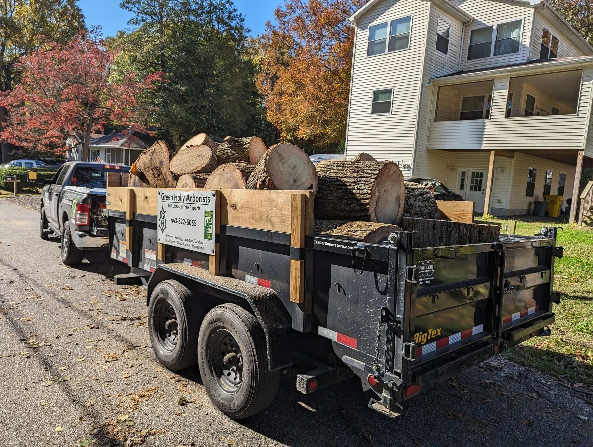 Black trailer piled with cut logs and rocks on a leafy residential street by houses