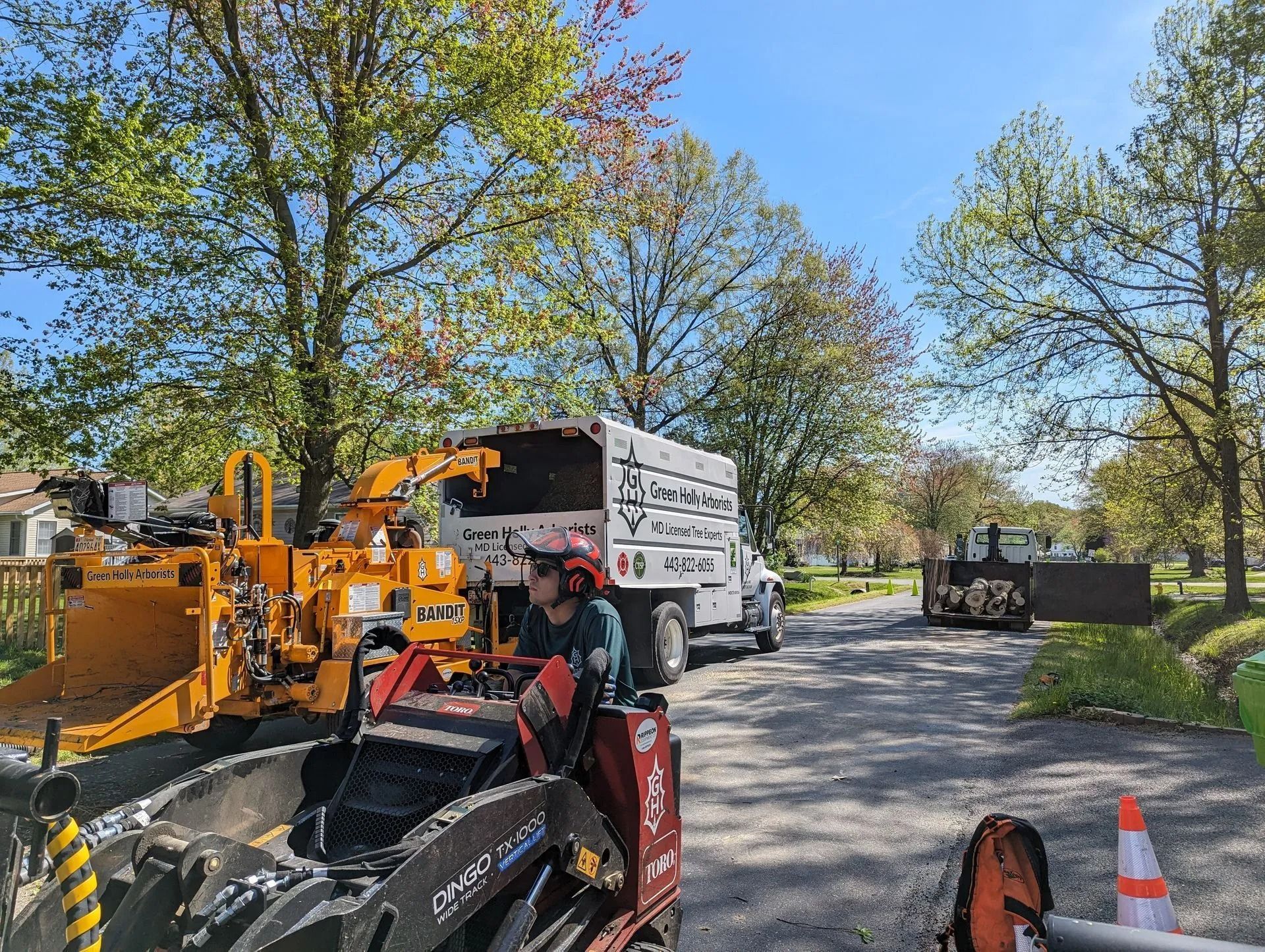 Roadwork scene with a white dump truck, orange paving machine, and worker in a green tree-lined street