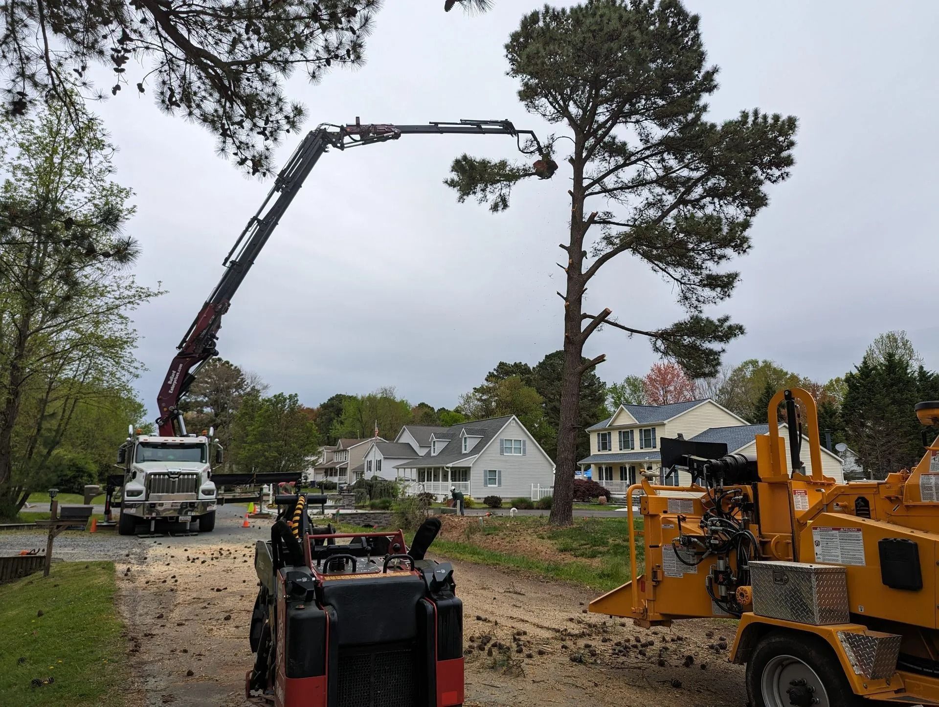 Construction crew and equipment clearing a roadside with cranes, trucks, and tree work near houses