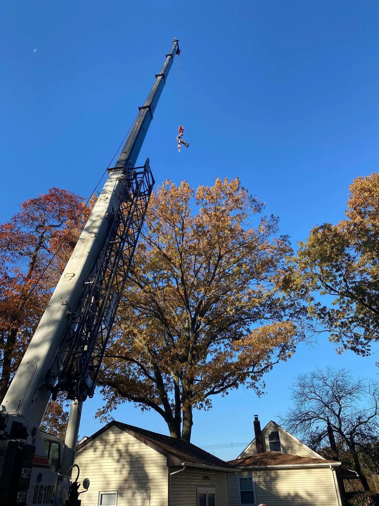 Cherry picker lifting a worker above a house amid autumn trees under a clear blue sky