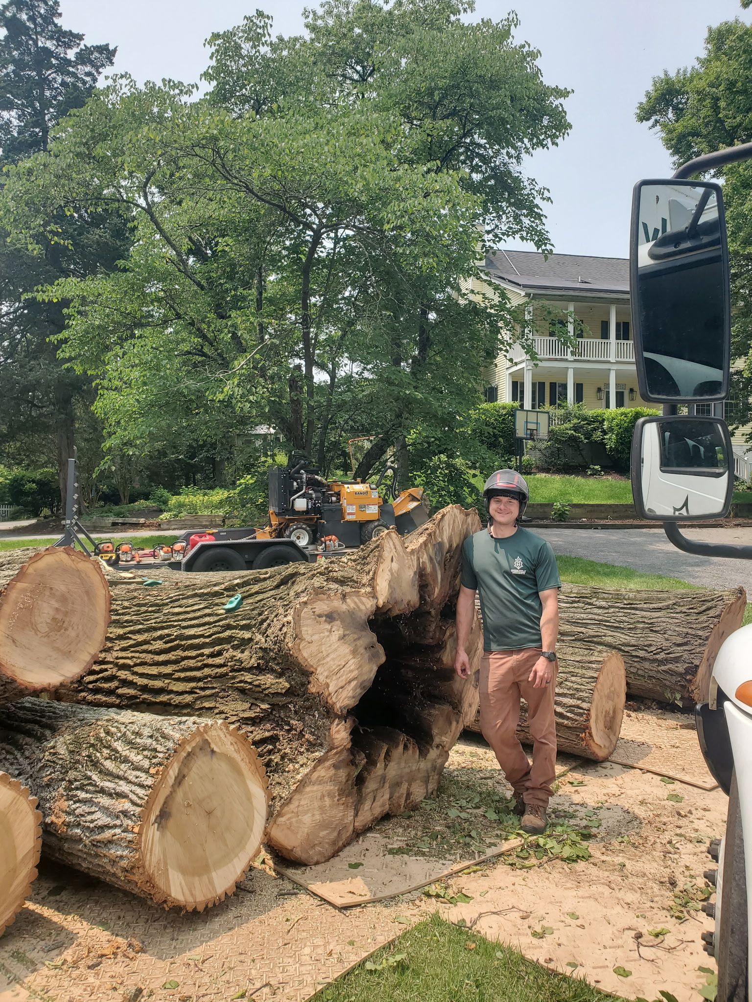 Person standing beside stacked cut logs and a wood chipper in a wooded yard