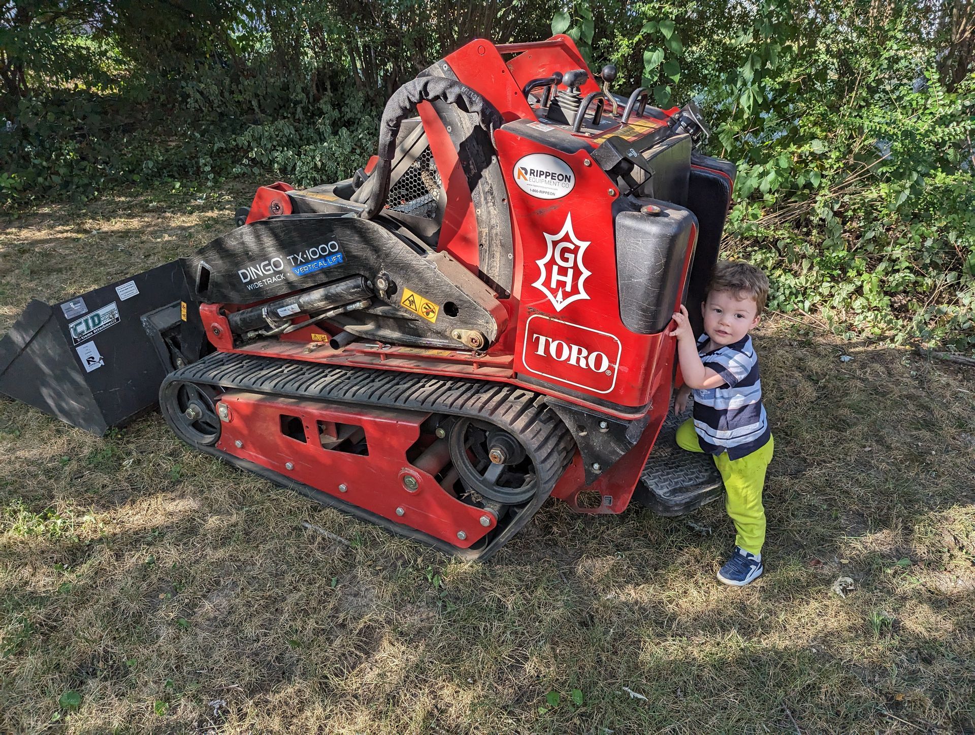 Child standing beside a red TORO tracked machine in a grassy wooded area