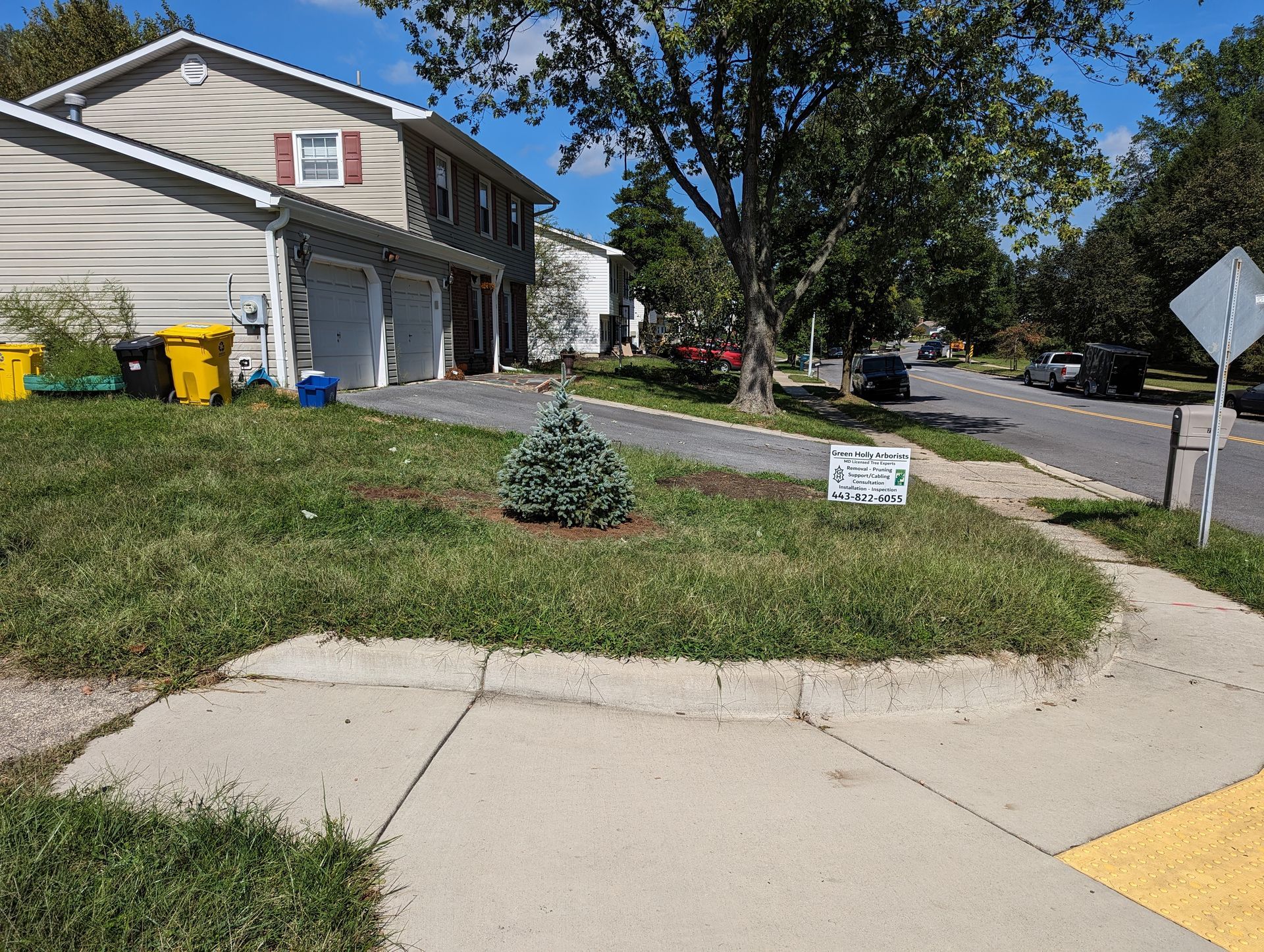 Suburban house at a street corner with a sloped lawn, trees, and trash bins by the garage.