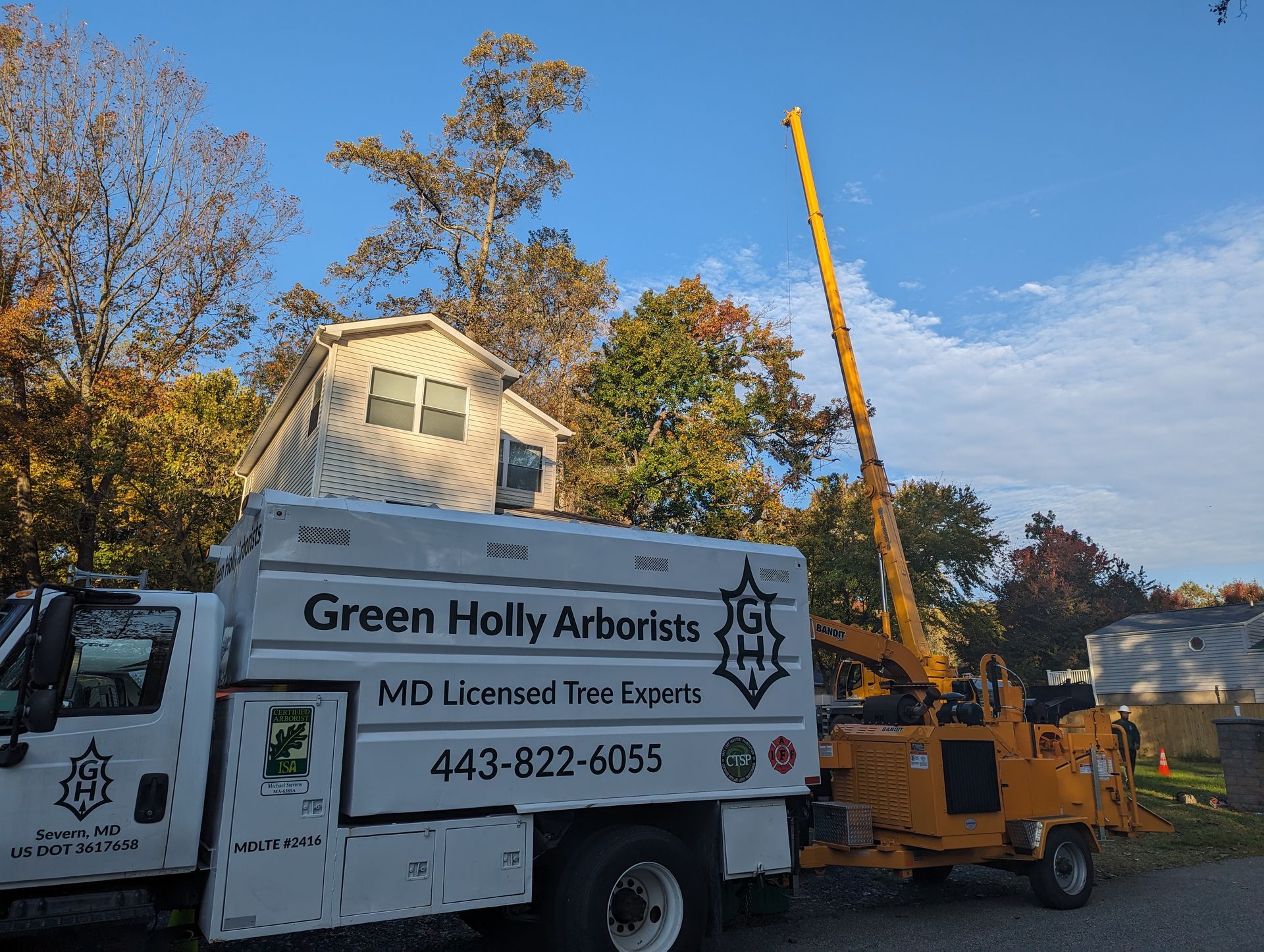 Green Holly Arborists truck and yellow tree service lift parked by a house in autumn.