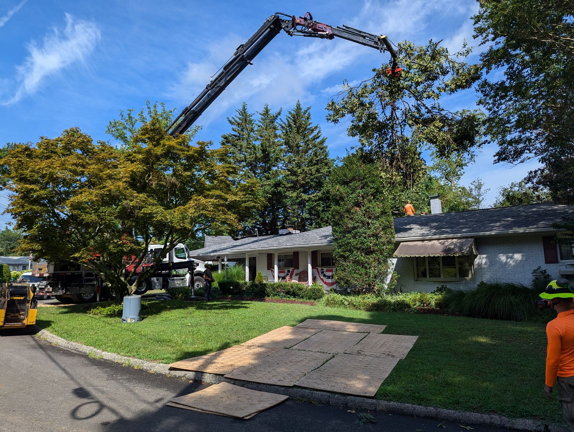 Tree-trimming crane over a suburban house, removing branches beside a leafy front yard.