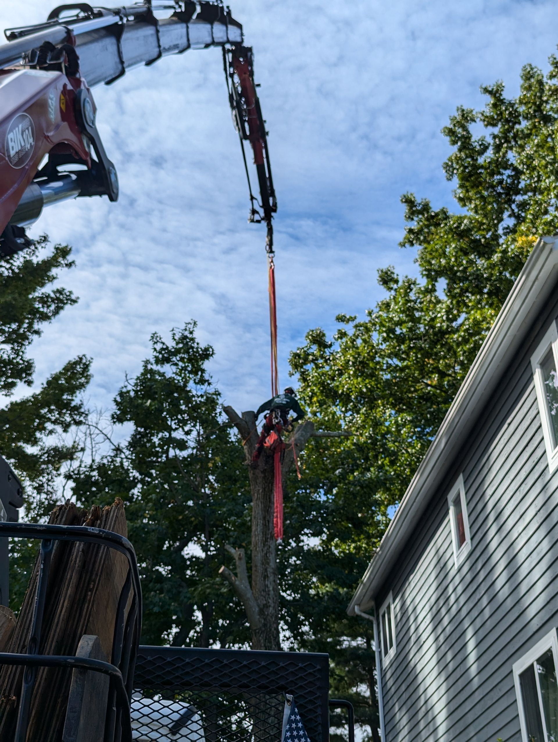 Worker hanging from a crane boom above a house roof, with trees and blue sky in the background