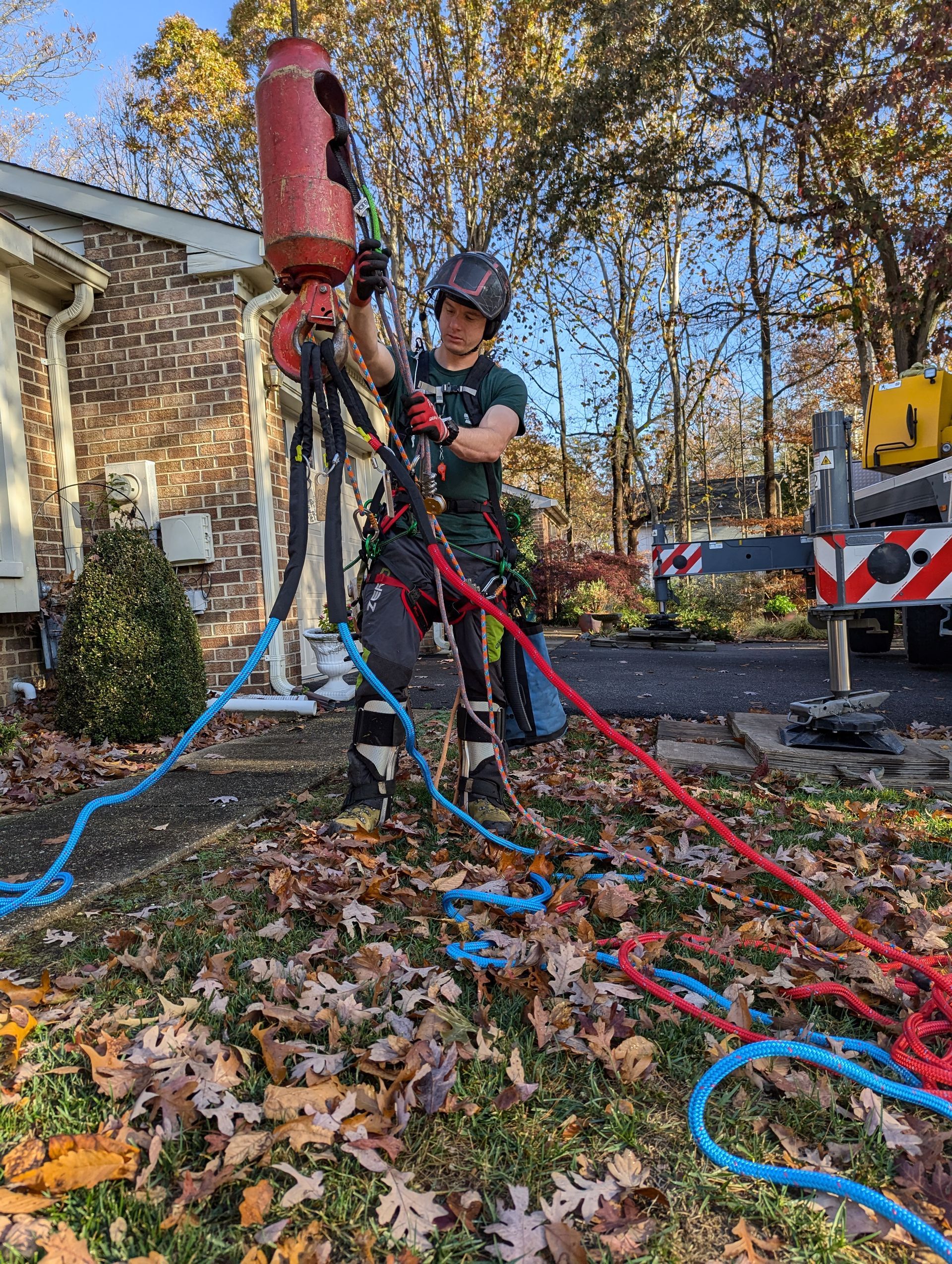 Worker operating a red hydraulic lift with blue hoses beside a stone house in a leafy yard