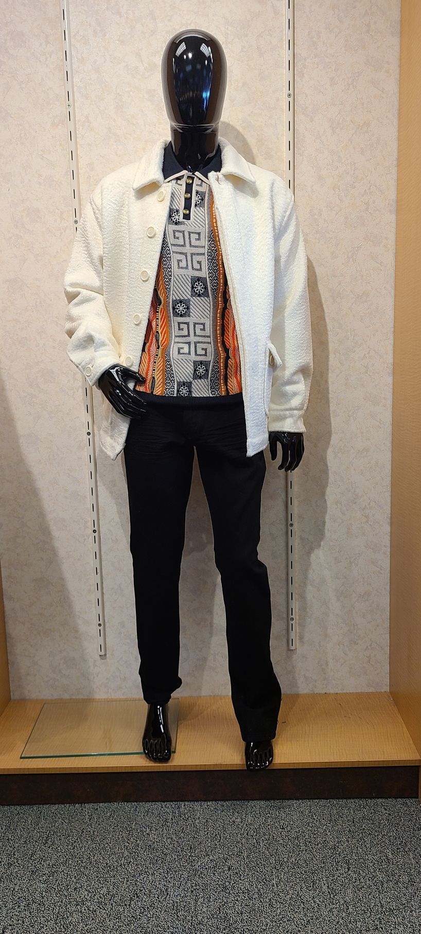 Mannequin wearing a white jacket, patterned shirt, black pants, and black shoes against a beige wall.