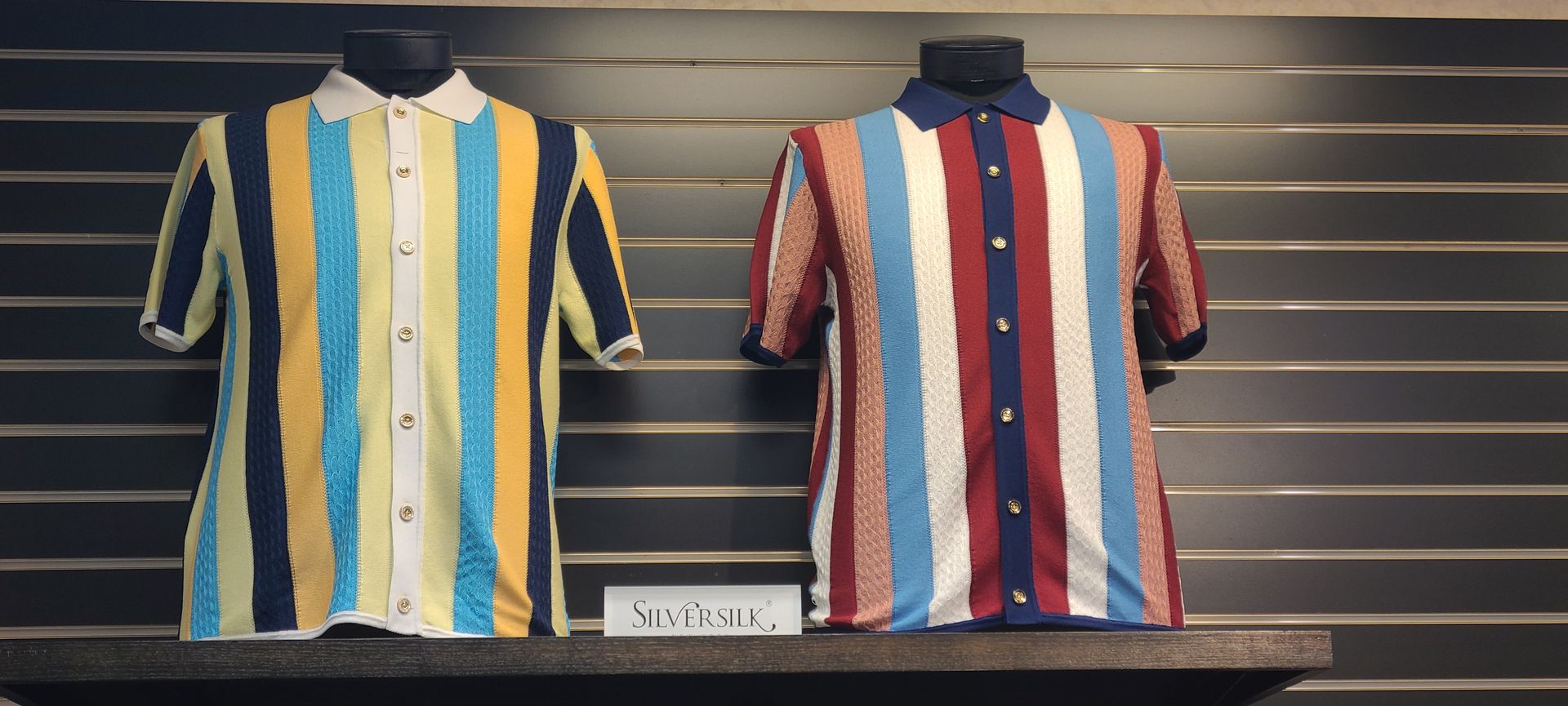 Two striped short-sleeve shirts are on display, one yellow and blue, one red and blue, hanging on a wall.