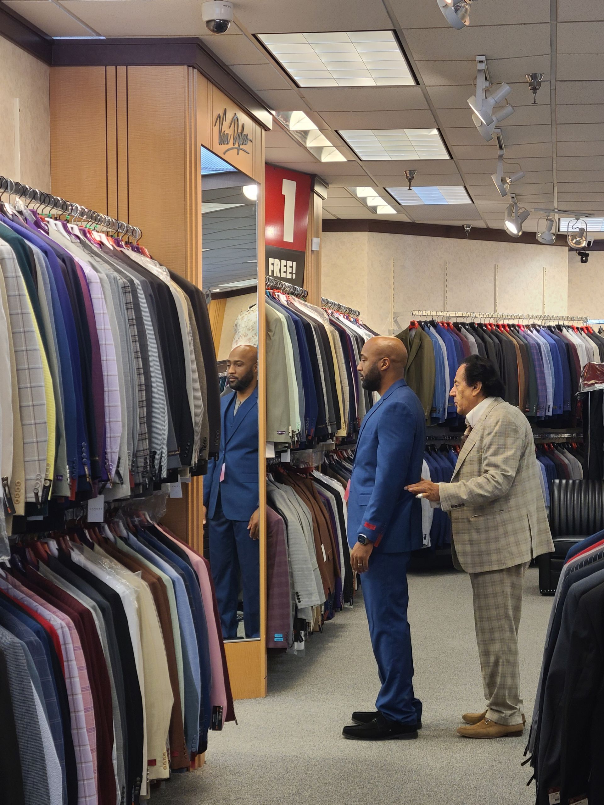 People browse suits in a clothing store aisle, with racks of jackets and dress shirts on both sides.