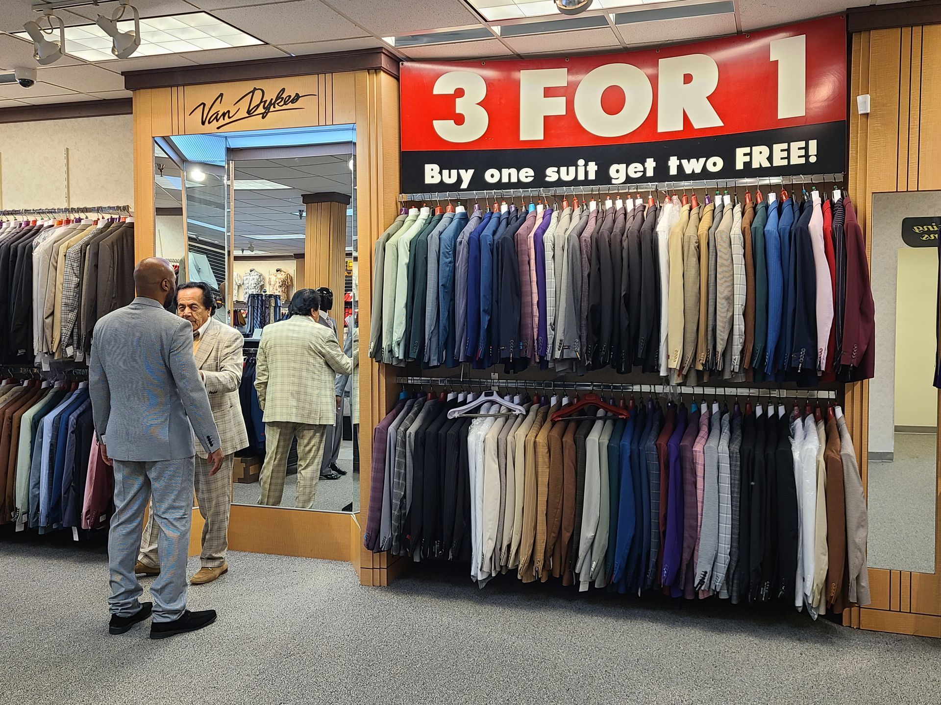 Suit store sale display with 3 FOR 1 sign and rows of hanging suits in a mall aisle
