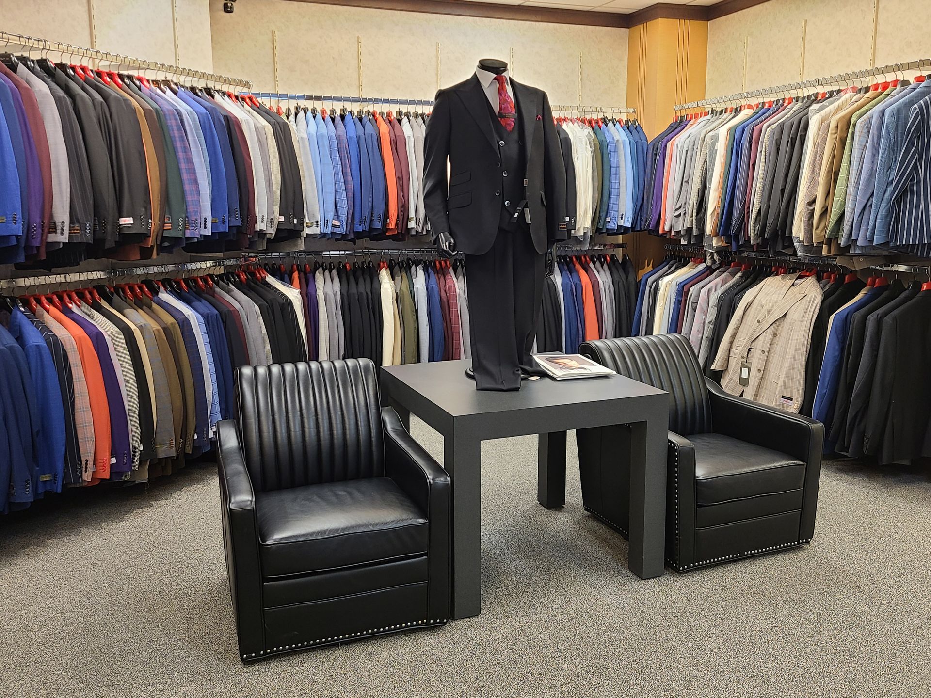 Men’s clothing store with suits on racks and a black suit displayed on a table between two chairs.
