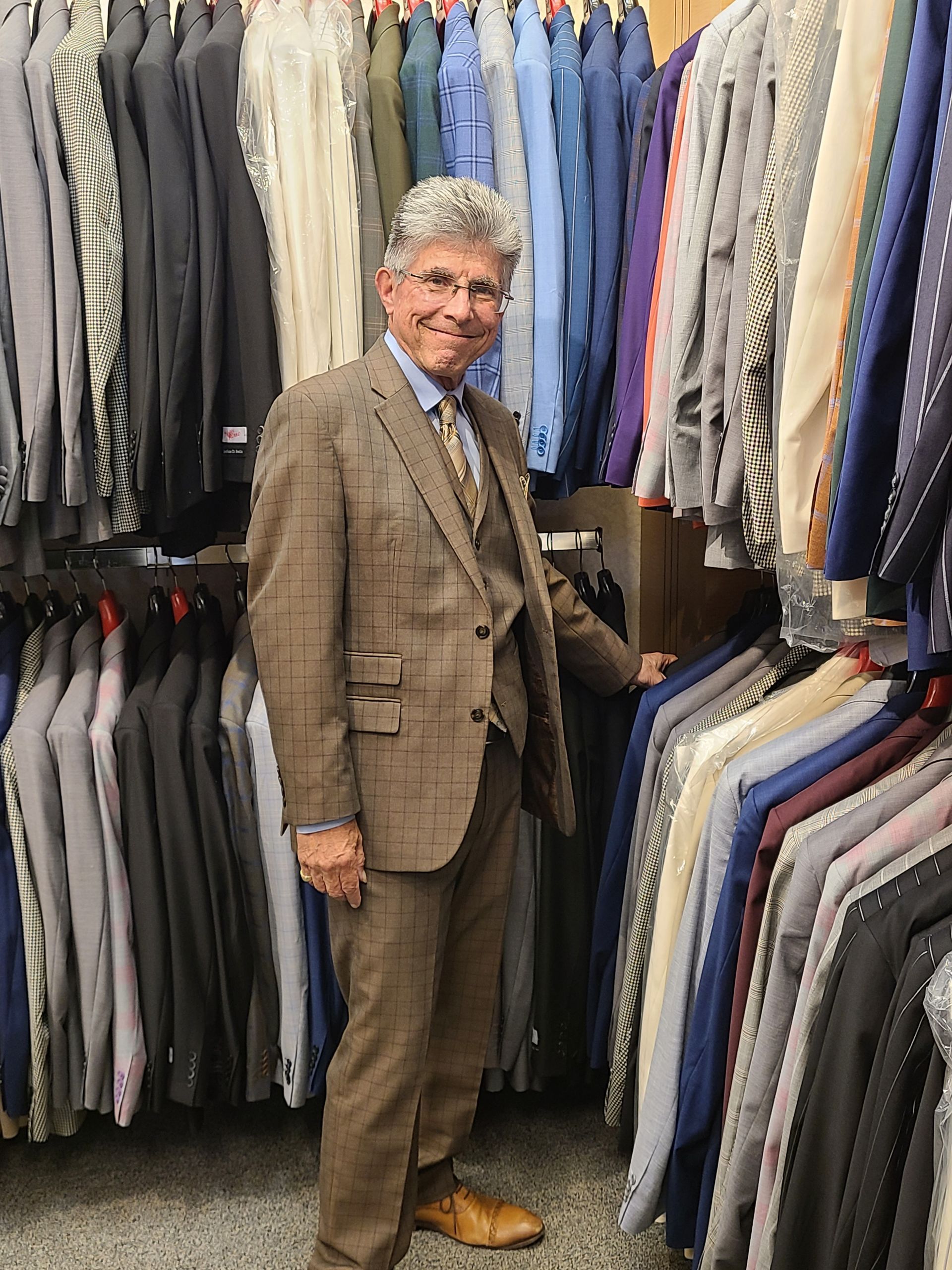 Man in a brown suit standing in a clothing store aisle between rows of hanging jackets and suits