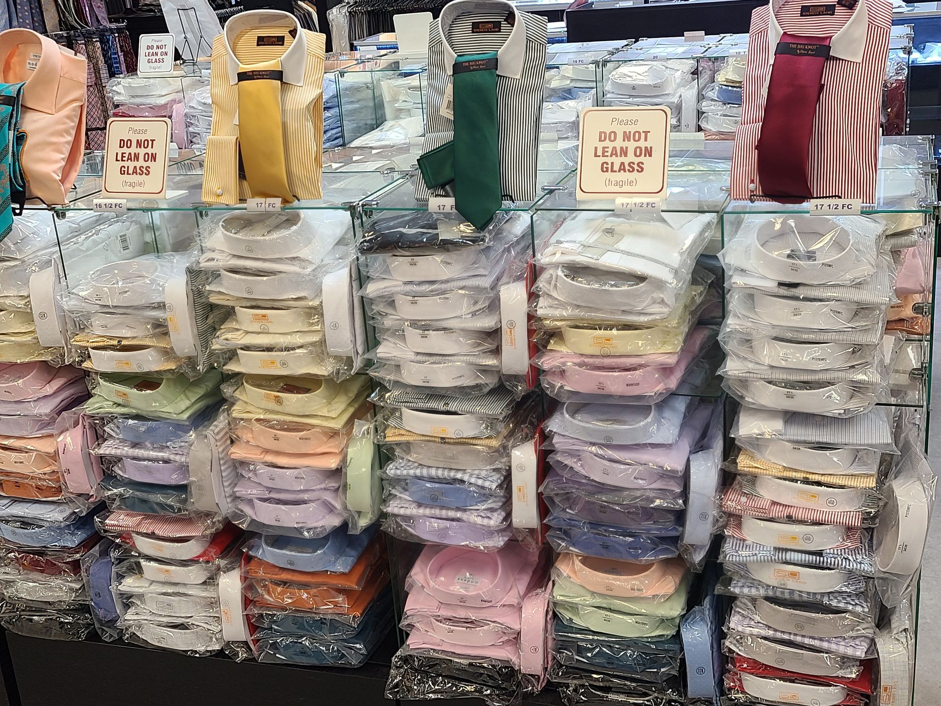 Stacks of packaged shirts in assorted colors are displayed at a market stall, with folded bags hanging above.