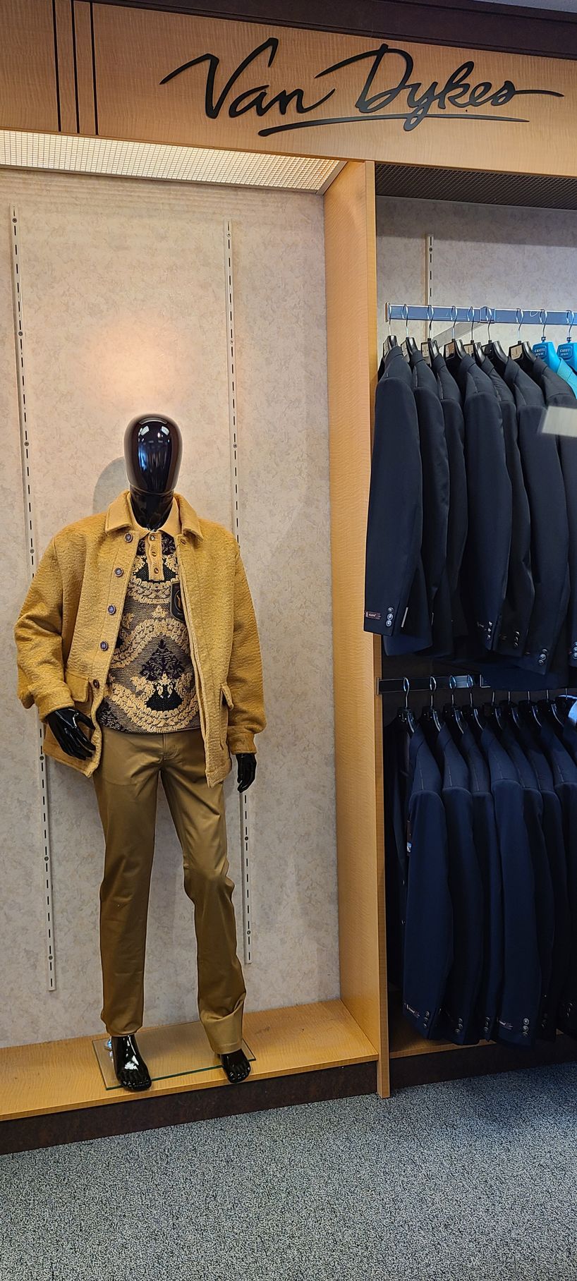 Mannequin in a tan jacket and pants beside a rack of dark suits in a clothing store.
