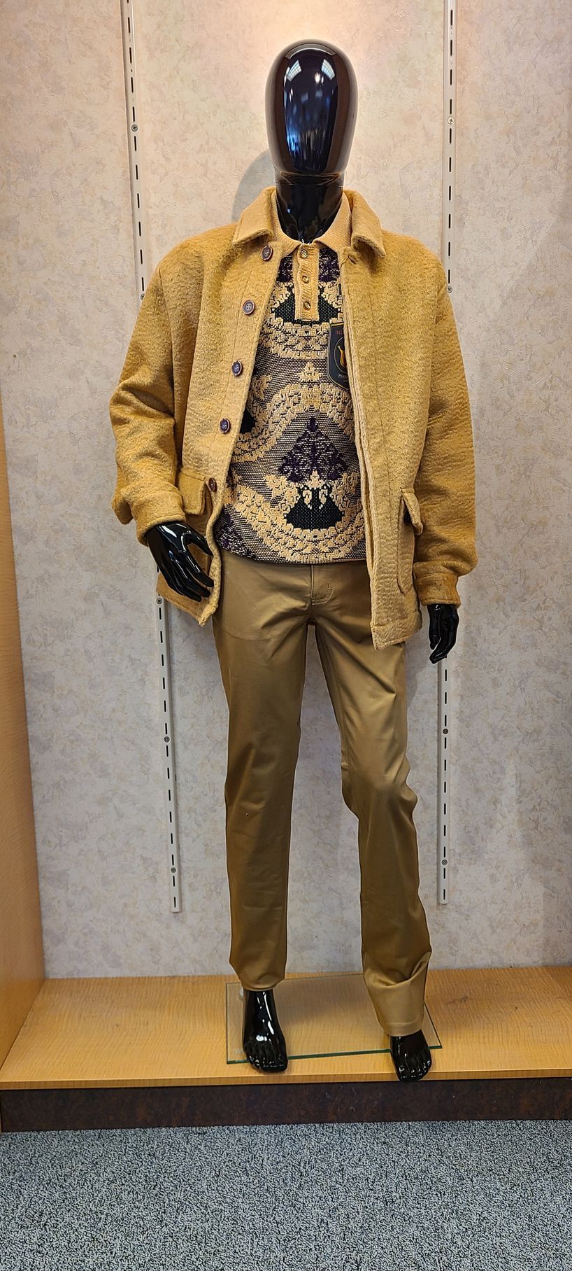 Mannequin in yellow jacket, patterned shirt, tan pants, and black shoes against a textured wall.