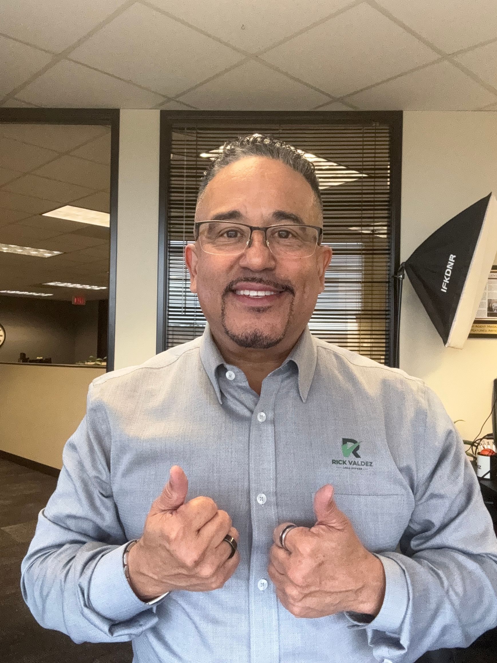 A man wearing glasses and a grey shirt is giving a thumbs up.