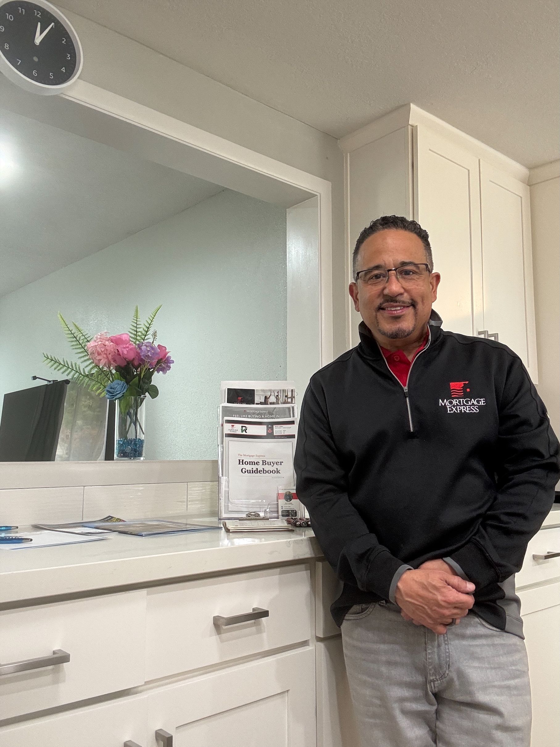 Man in black jacket leans on kitchen counter. White cabinets, flowers, clock in background.