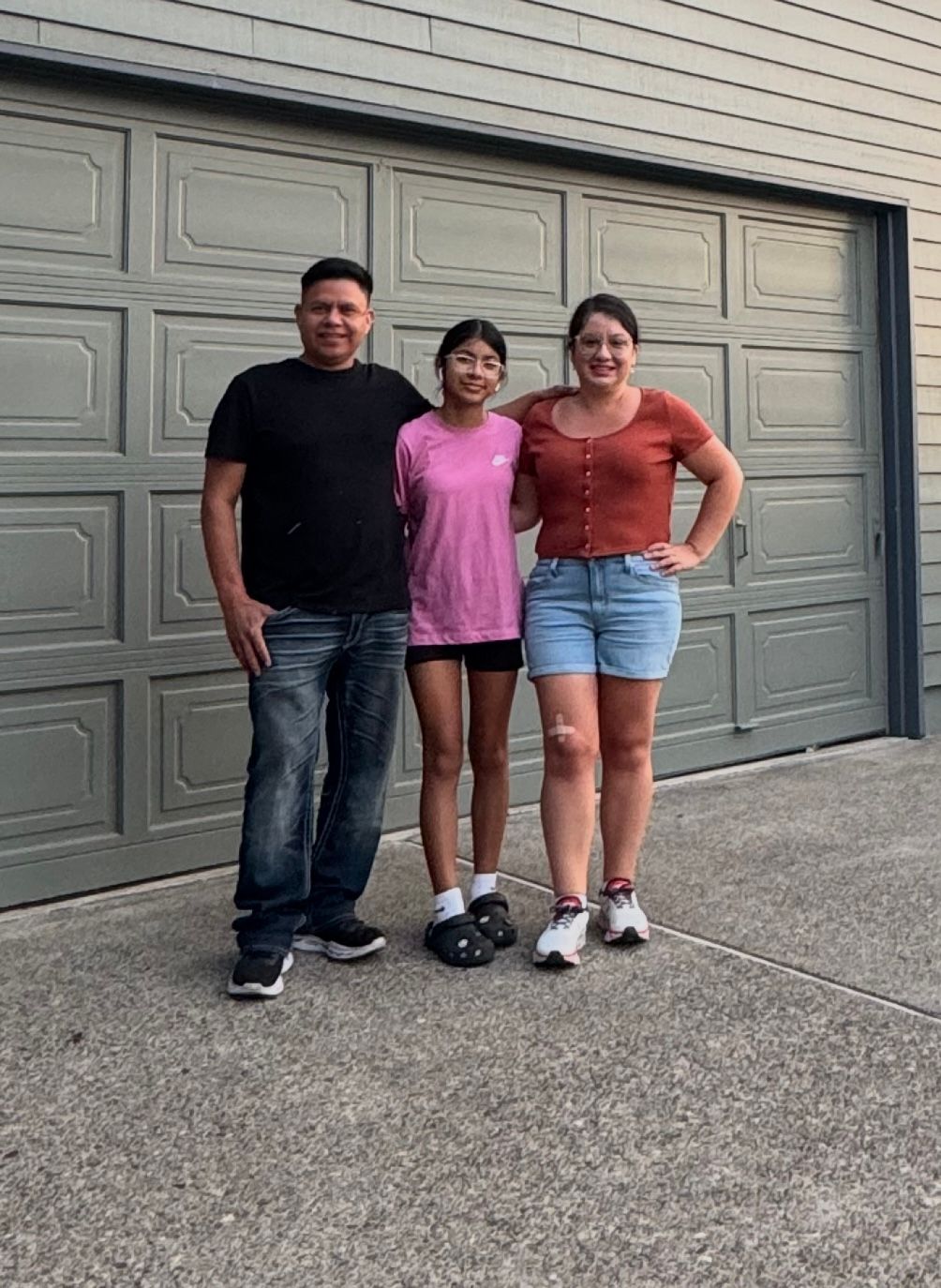 Three people pose in front of a gray garage door: a man in black, a girl in pink, and a woman in orange.