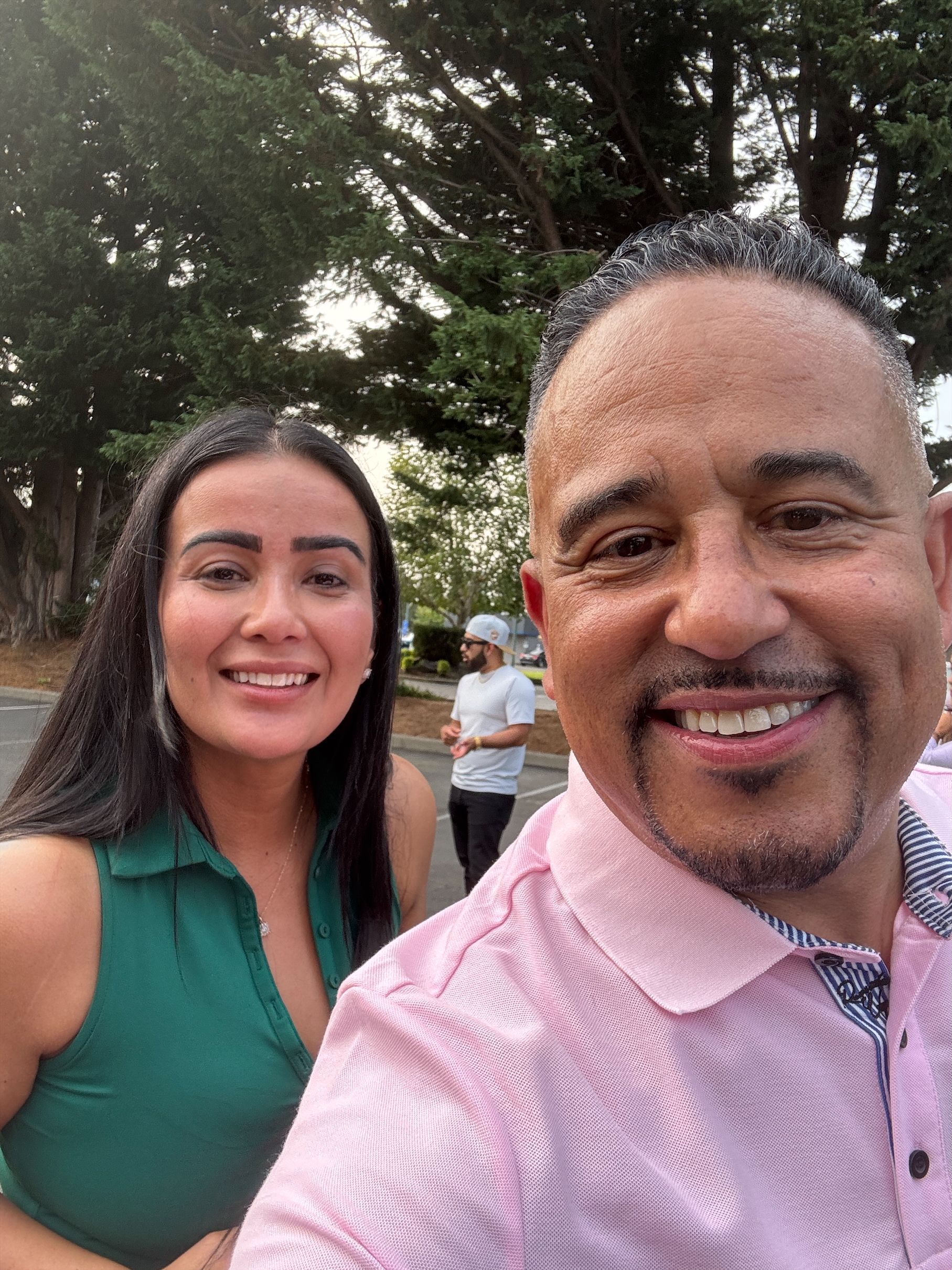 A man and woman smiling for a selfie outdoors. The man wears a pink shirt. The woman wears a green top.