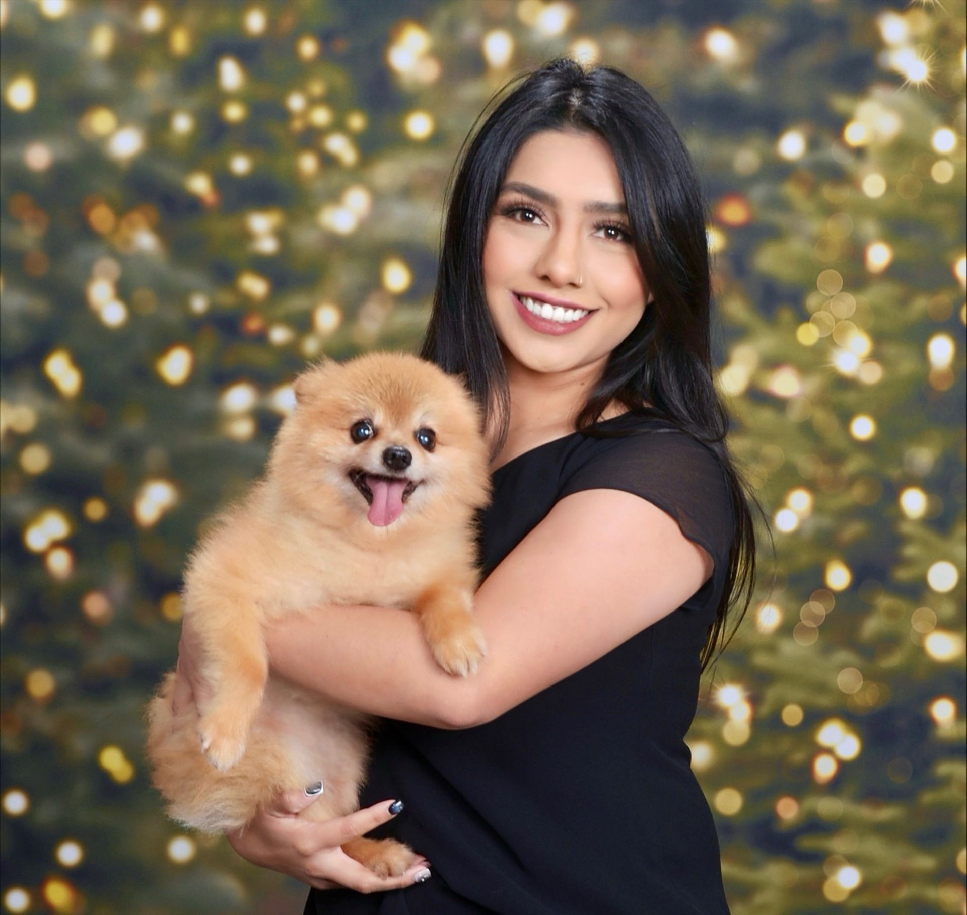 Woman in black dress holds a small, fluffy, tan dog in front of a blurred Christmas light background; both smiling.